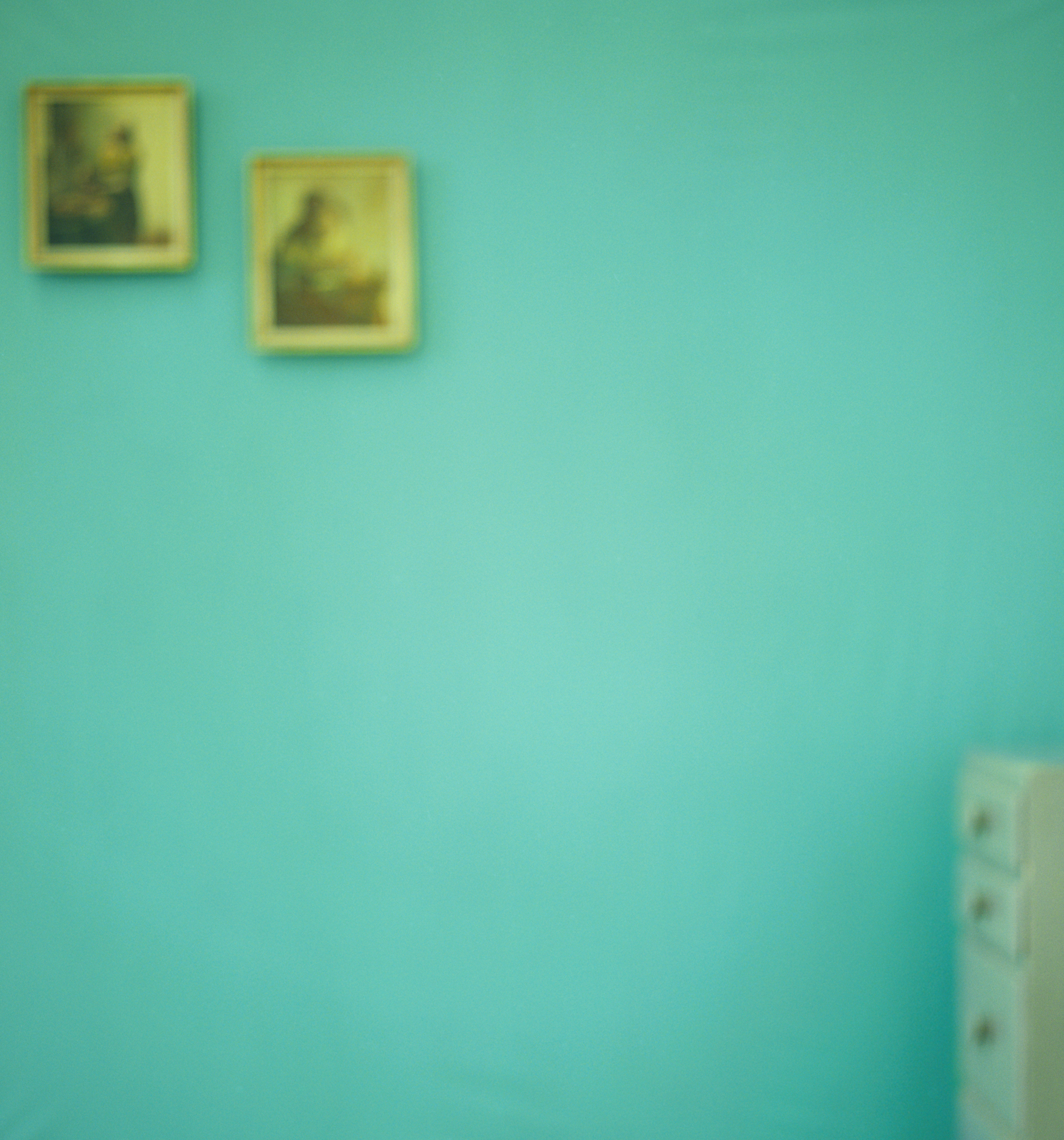 a photograph by Uta Barth showing a blurry greenish wall with two reproductions of Vermeer hanging on it and a small white dresser to the right